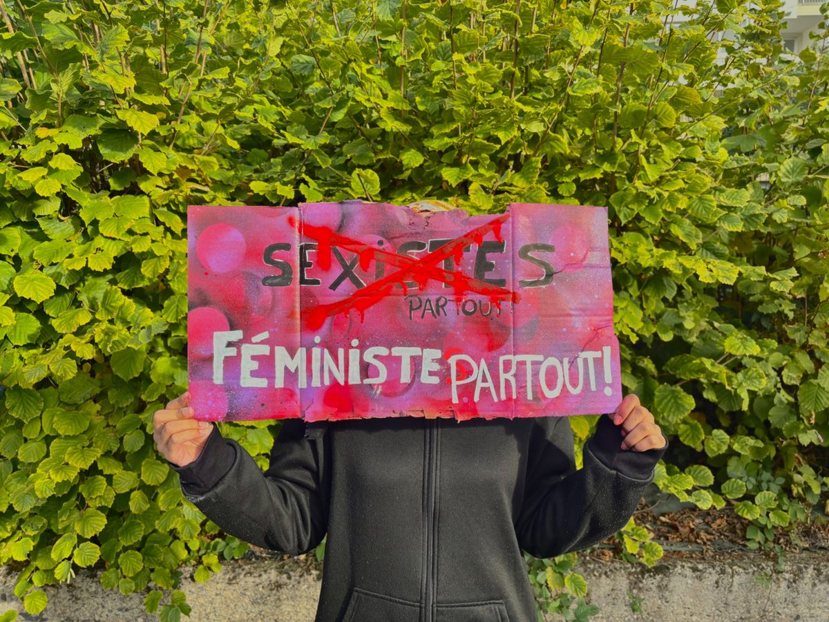 Pancarte rose et rouge tenue devant une haie : « Sexistes partout » barré, puis « Féministe partout ! »
