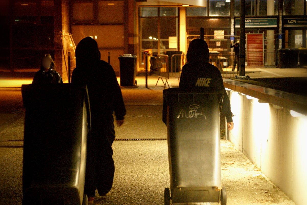 Deux silhouettes encapuchonnées poussant des poubelles de nuit devant un bâtiment de l'université