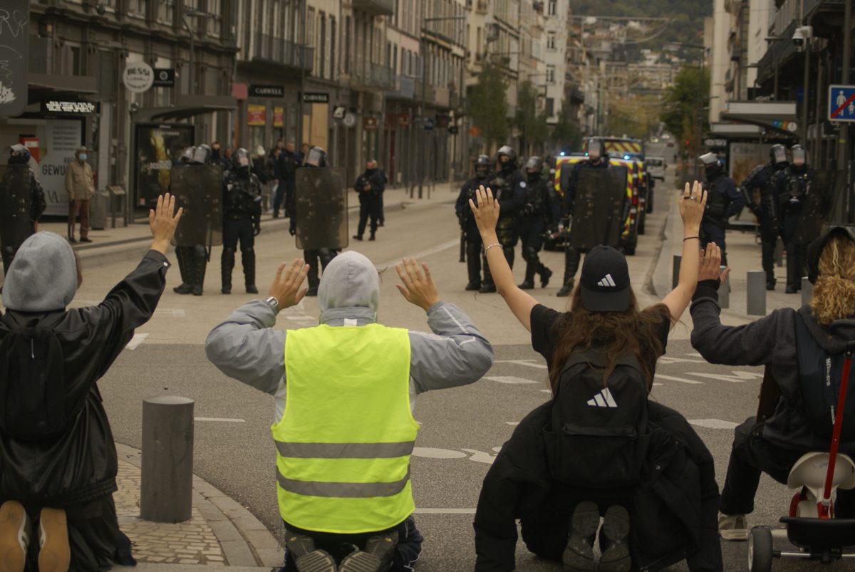 Manifestants à genoux, mains levées, face à une rangée de policiers casqués dans une rue du centre-ville