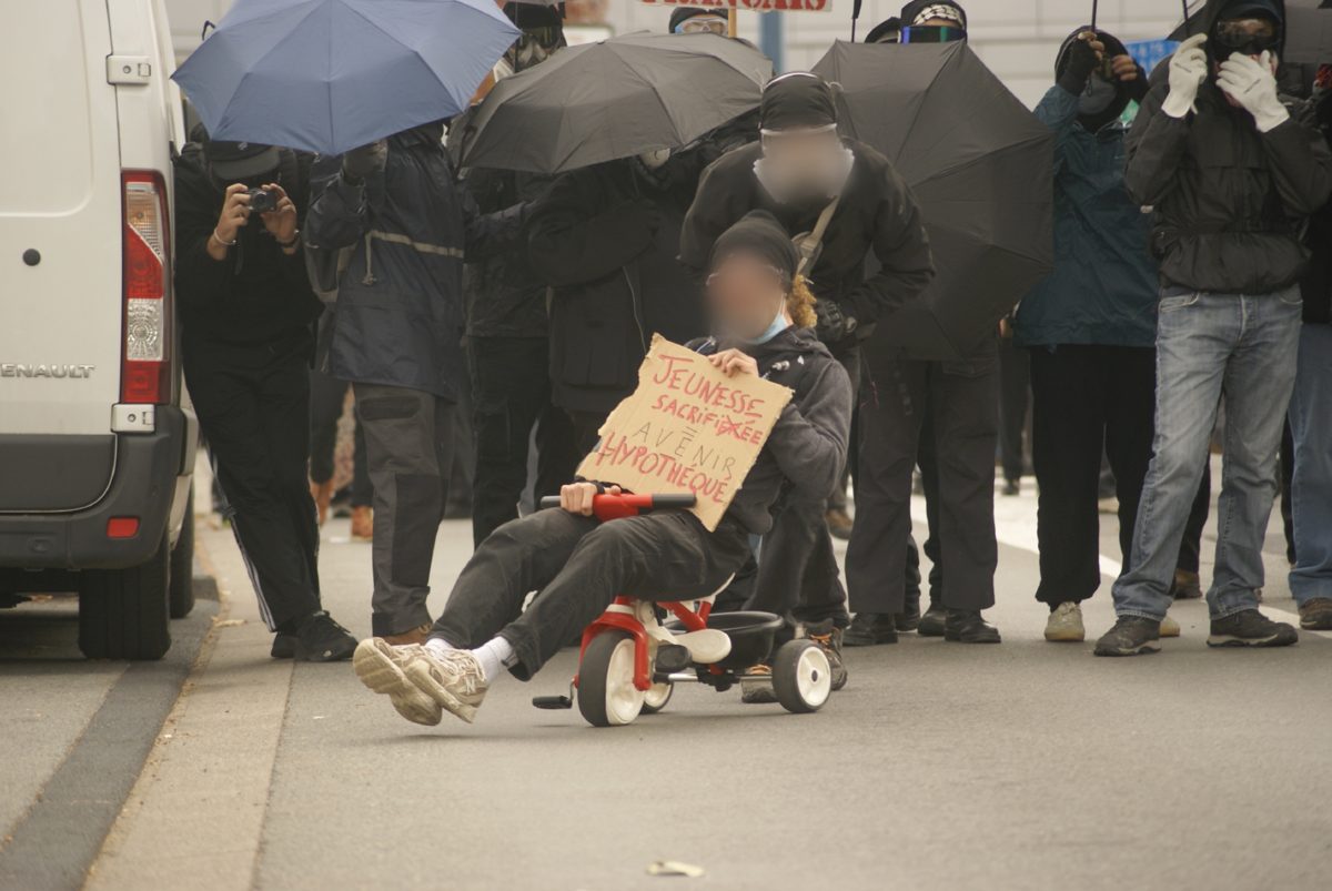 Personne assise sur un tricycle rouge tenant une pancarte « Jeunesse sacrifiée, avenir hypothéqué », entourée de manifestants sous des parapluies noirs